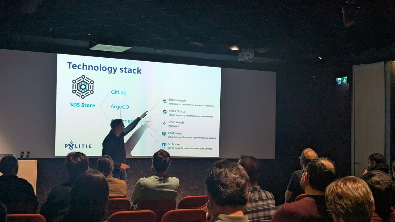 Politie speaker pointing at the SDS Store technology stack: GitLab, ArgoCD and Kubernetes with Elasticsearch, Kafka Strimzi, OpenSearch, PostgreSQL and S3