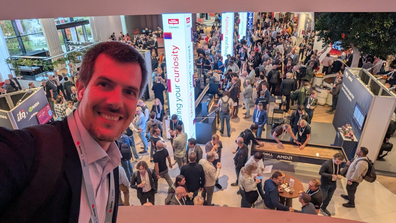 Selfie of Luca Berton on a balcony above the expo floor at Red Hat Summit Connect 2025, with Bring your curiosity banners, an ITQ and NVIDIA stand and a Dell and AMD stand below