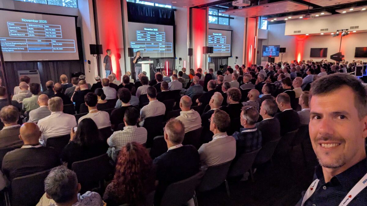 Luca Berton selfie at Red Hat Tech Day Netherlands 2026 keynote — packed room with red uplighting, November 2025 model comparison slides on screen