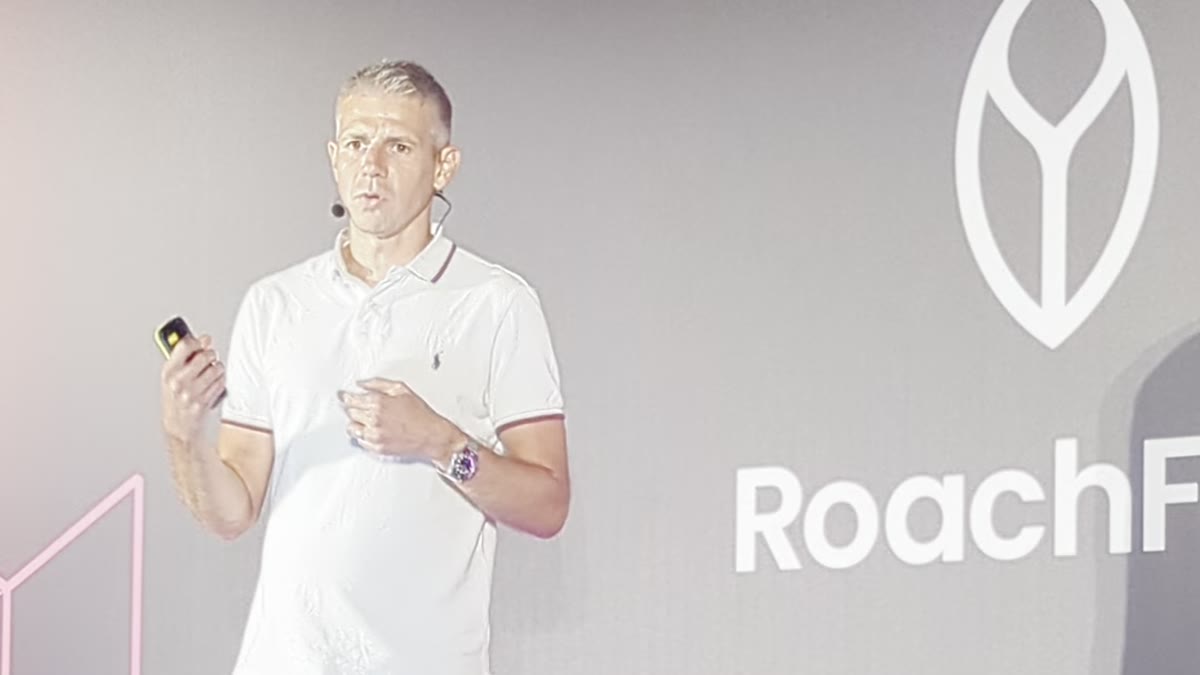 A speaker on stage in front of the RoachFest logo at RoachFest London 2026, mid-explanation of cross-cluster Kubernetes Pod Disruption Budgets