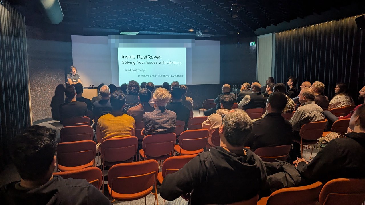 The Inside RustRover title slide with the speaker at the lectern and a full room in front of the screen