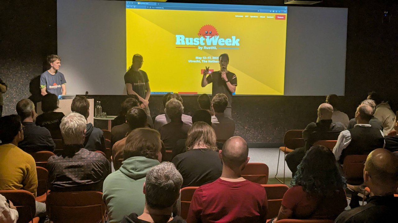 Speakers on stage in front of the yellow RustWeek by RustNL site, announcing 13–17 May 2025 in Utrecht