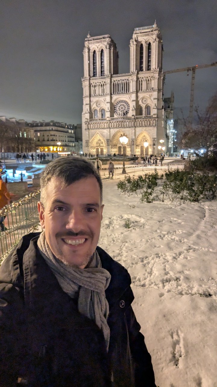 Luca smiling in a scarf in front of the west facade of Notre-Dame de Paris at night, with snow covering the ground of the square