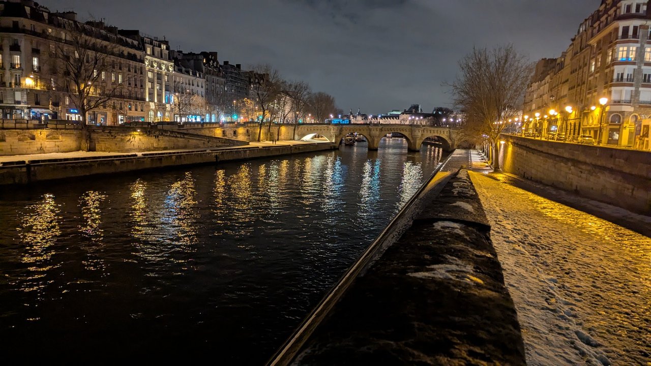 A quay along the Seine at night, the stone path covered in snow and lit by street lamps, with a bridge and the lights of Paris reflected in the river