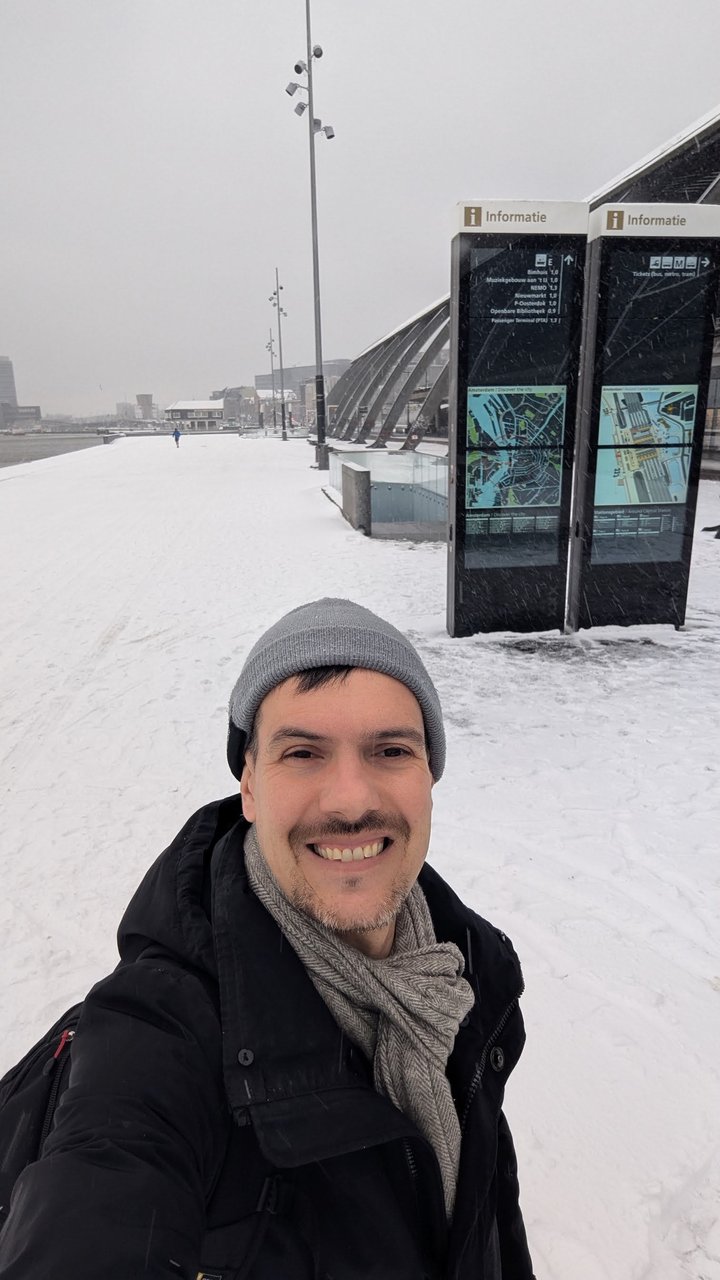 Luca in a grey beanie and scarf, smiling, on a snow-covered waterfront promenade with information signs behind him