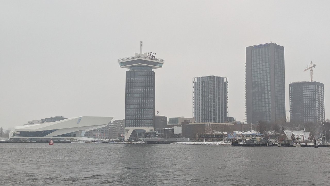 The Amsterdam IJ waterfront under a grey winter sky, with the A'DAM Tower, the EYE Filmmuseum and snow on the low roofs along the water