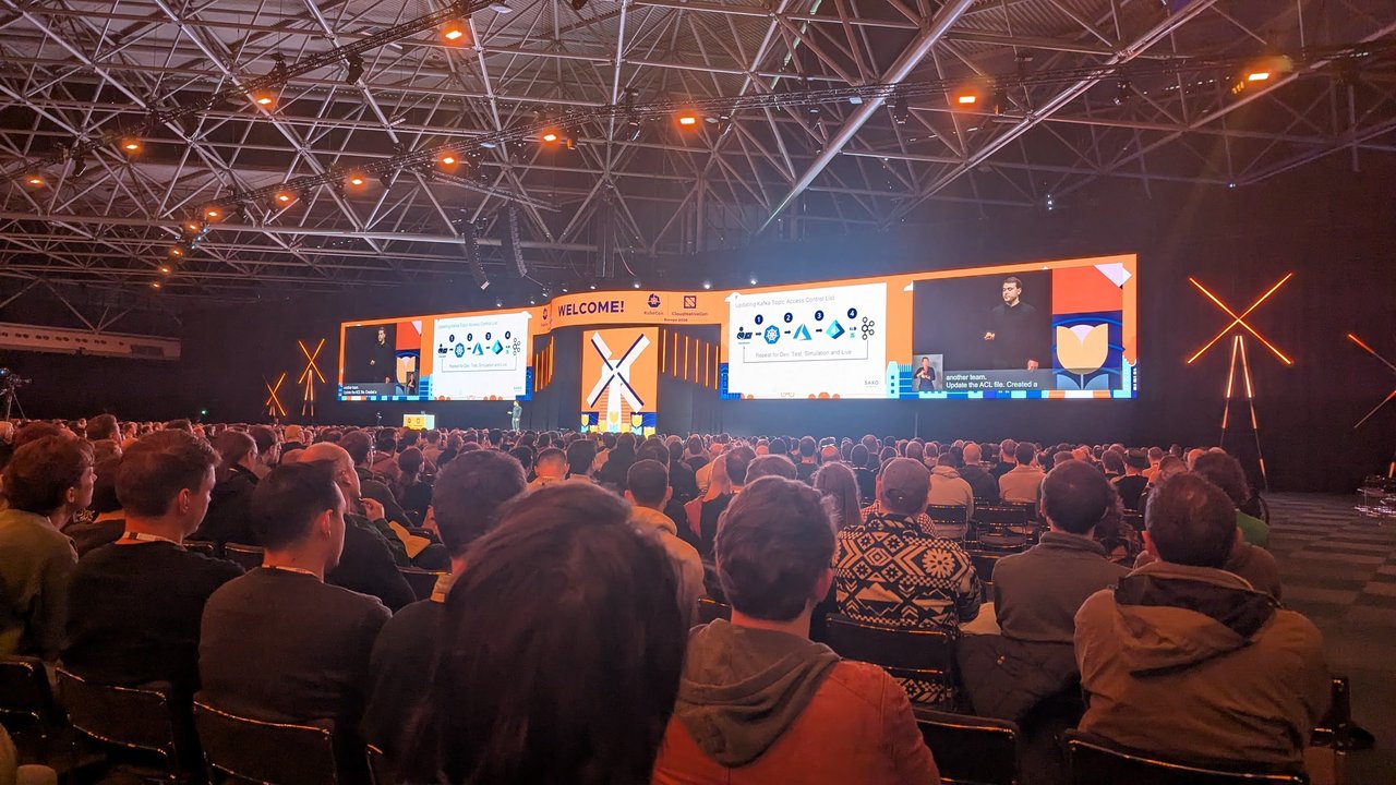 The KubeCon Europe 2026 keynote hall in Amsterdam, with the Saxo slide Updating Kafka Topic Access Control List on both stage screens and a full audience in front