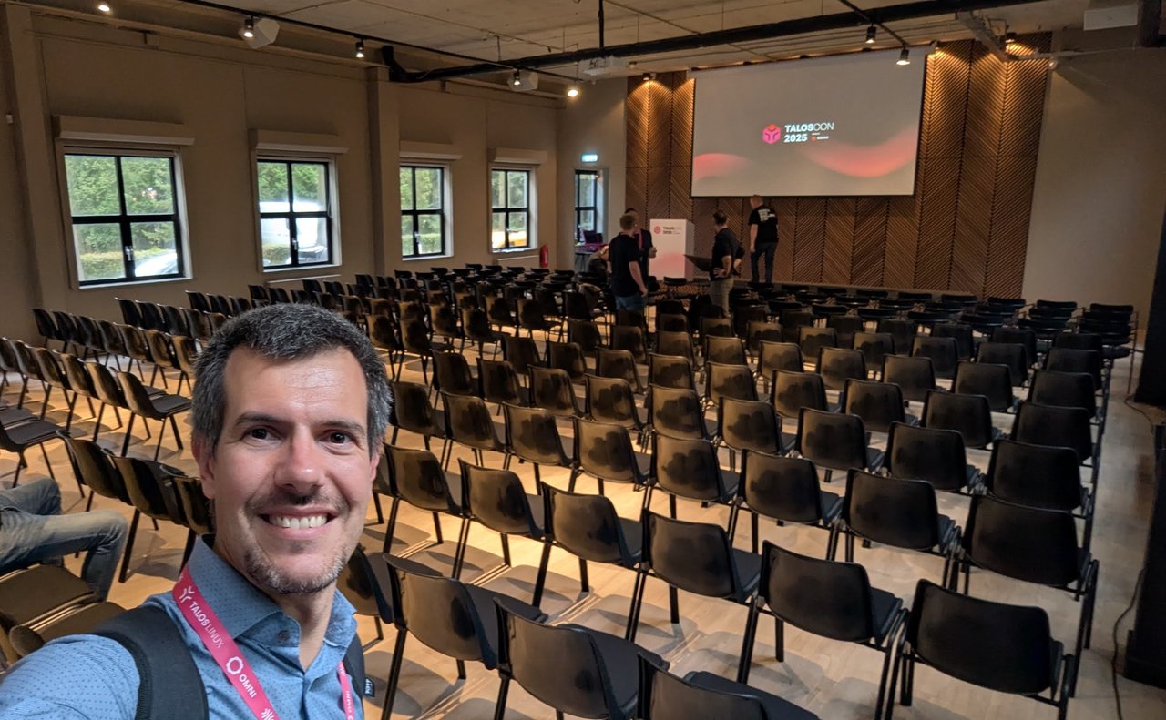 Luca Berton taking a selfie in the empty TalosCon 2025 plenary room, rows of black chairs and the TalosCon 2025 slide on the screen