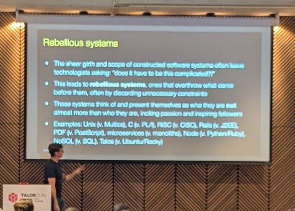 Keynote slide titled Rebellious systems with examples such as Unix versus Multics, RISC versus CISC, microservices versus monoliths and Talos versus Ubuntu or Rocky