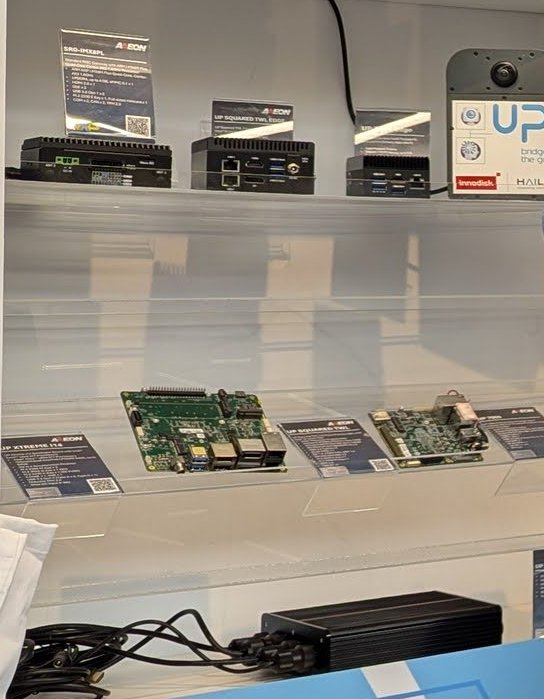 A glass display at the AAEON booth with compact fanless boxes and bare x86 boards, labelled with the UP brand