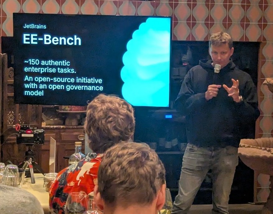 JetBrains EE-Bench slide at the evening session: about 150 authentic enterprise tasks, an open-source initiative with an open governance model, with the speaker beside the screen
