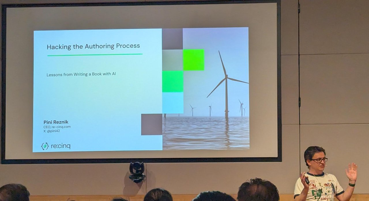 Pini Reznik on stage next to the title slide Hacking the Authoring Process: Lessons from Writing a Book with AI, with his name, CEO re-cinq.com and an offshore wind farm image