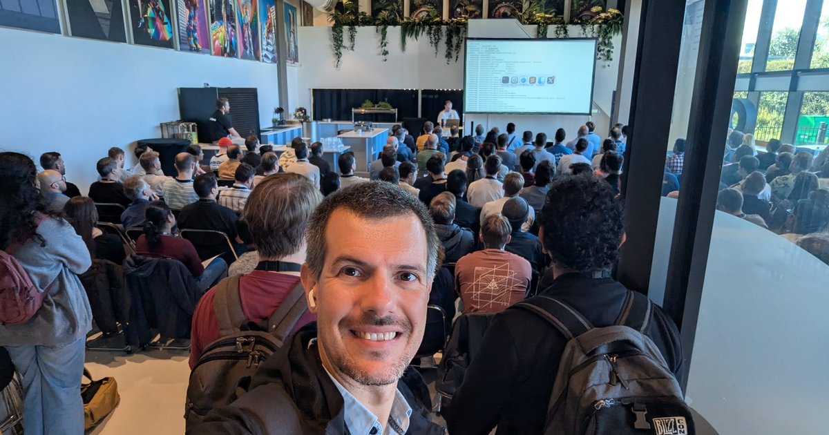 Luca Berton taking a selfie at the back of a packed keynote room at AI4Devs Amsterdam 2025, with live code on the projector screen