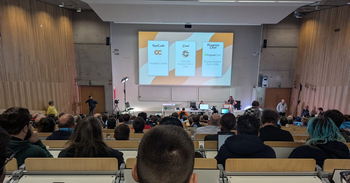 A full lecture hall at CfgMgmtCamp 2025 in Ghent with a slide showing Chef's history from OpsCode in 2008 to Progress Chef in 2020