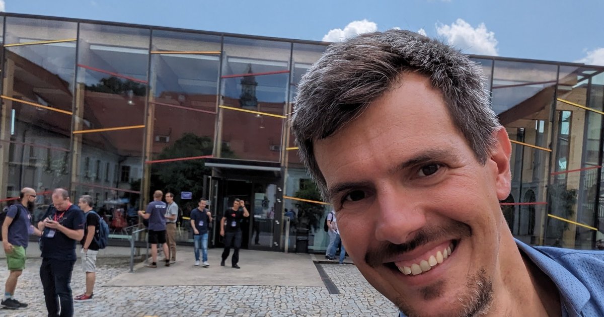 Luca Berton in the DevConf.cz 2023 venue courtyard in Brno, with attendees in front of the glass building