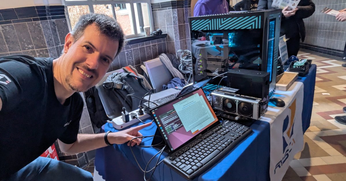 Luca Berton at a RISC-V stand at FOSDEM 2026, pointing at a laptop next to a large tower PC with a glass side panel