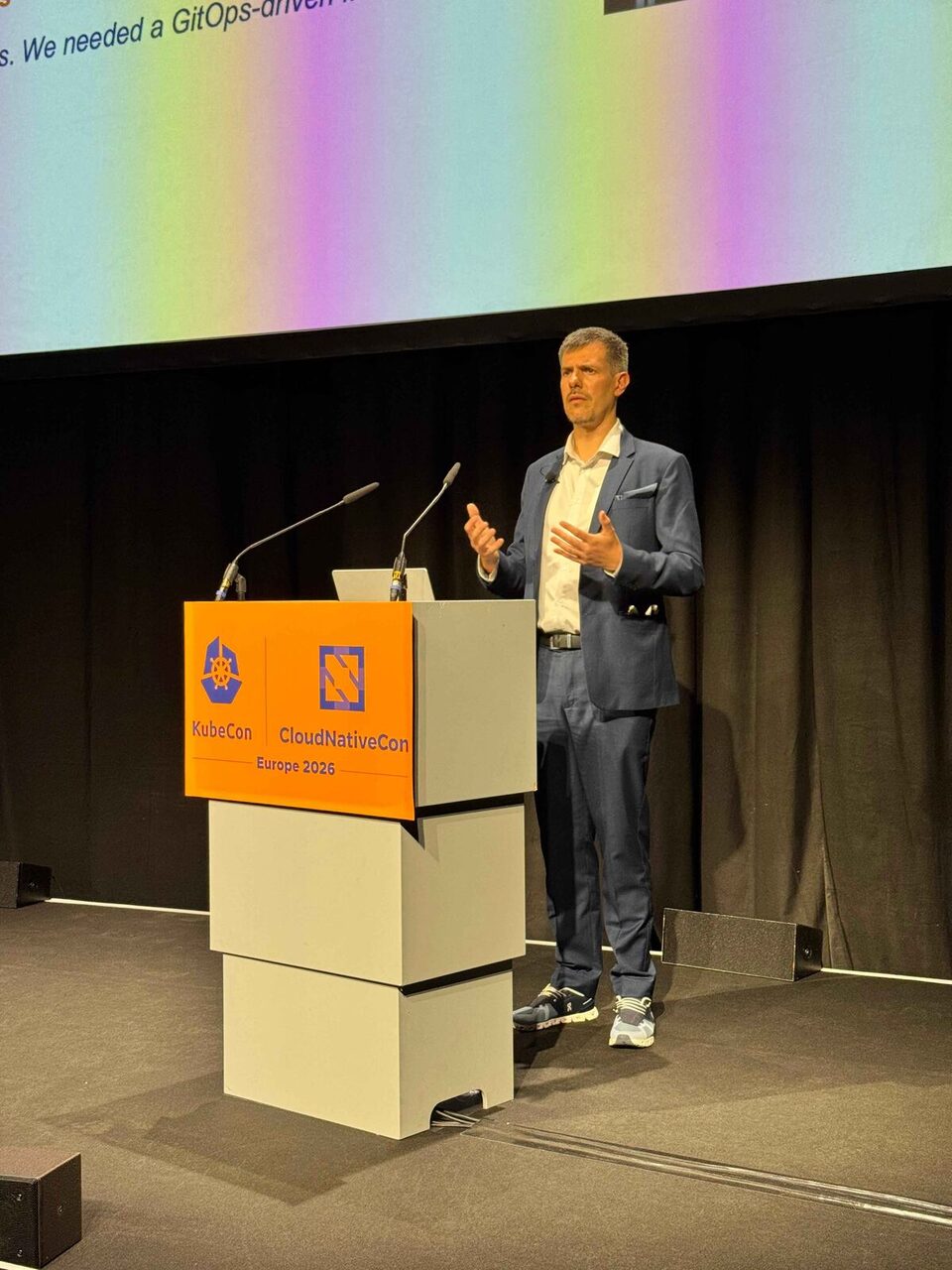 Luca Berton speaking at the podium, explaining the need for a GitOps-driven framework
