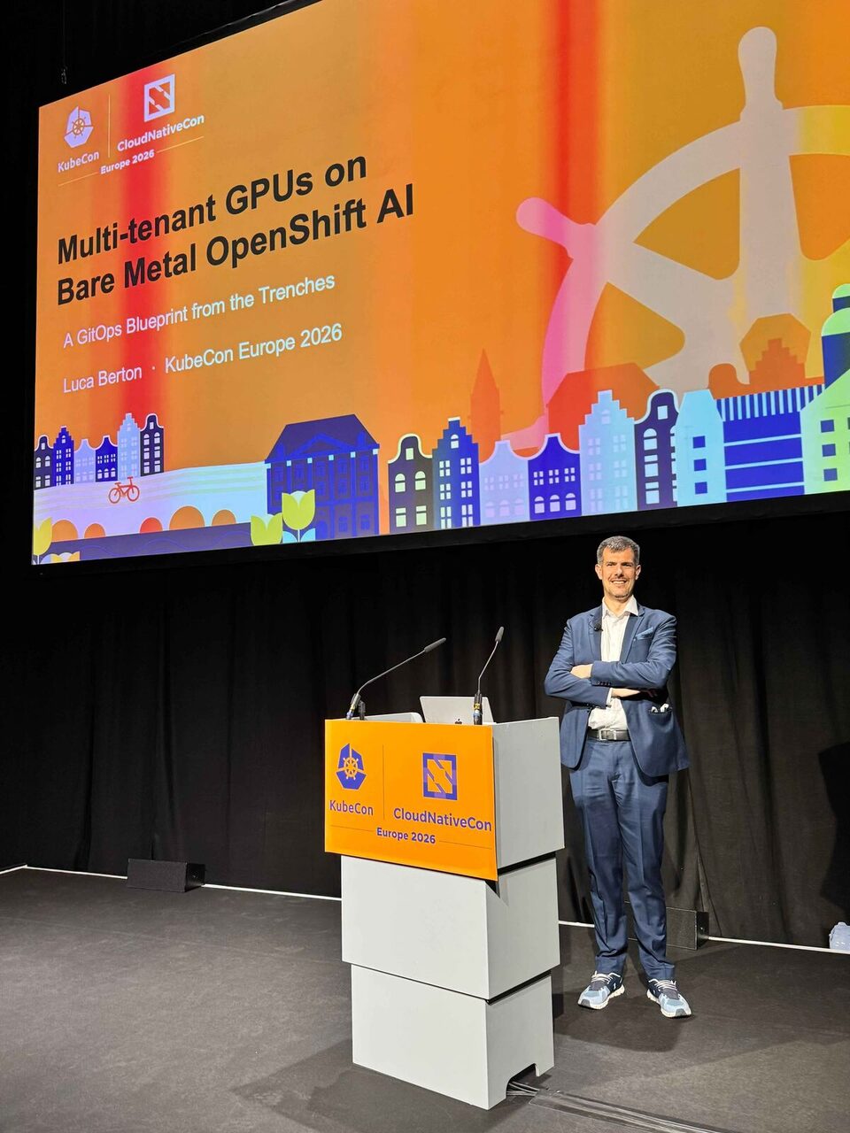 Wide shot of the KubeCon stage with the presentation title and Amsterdam-themed graphics