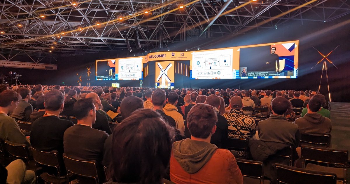 The KubeCon + CloudNativeCon Europe 2026 keynote hall at RAI Amsterdam, with a Saxo architecture slide on the big screens above a full audience