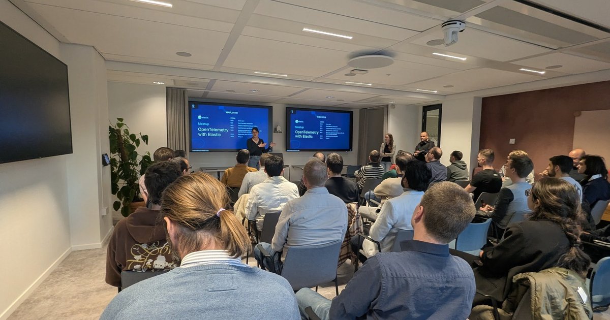 Elastic presenter at the OpenTelemetry with Elastic meetup in Amsterdam, with the welcome and agenda slide on two screens and the audience seated