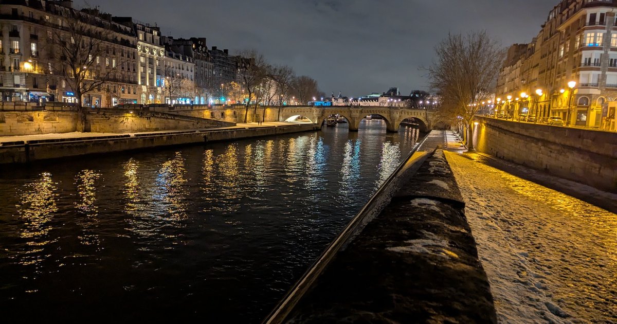 A snowy quay along the Seine in Paris at night, with lit buildings and a stone bridge reflected in the river