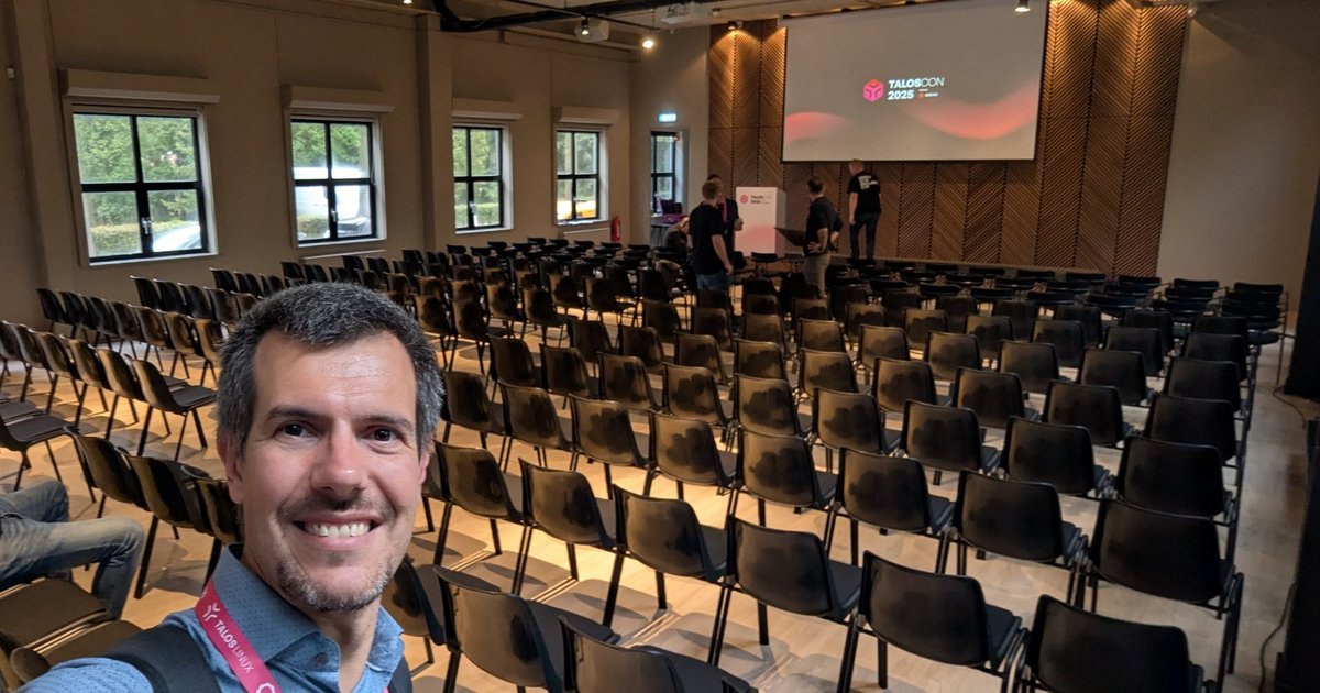 Luca Berton in the empty TalosCon 2025 plenary room at Venue Collective in Amsterdam, with the TalosCon 2025 slide on the screen behind him